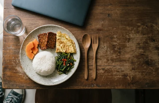 Sepiring hidangan nasi dengan tempe goreng, telur dadar, tumis kangkung, dan potongan pepaya di meja kayu.
